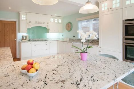 A general interior view of a white fitted shaker style kitchen with island, wicker bar stools, glass shade ceiling lights, potted orchid and fruit bowl. Credit : getty